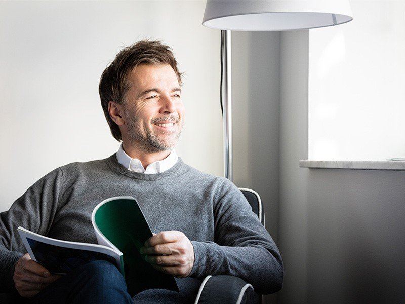 Life Extension, smiling middle-aged man sitting in a chair with an open magazine, in a grey sweater next to the window